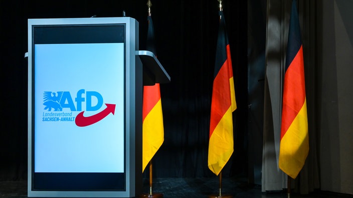 Das AfD-Logo am Rednerpult mit Deutschlandfahnen im Hintergrund auf der Bühne im Versammlungssaal