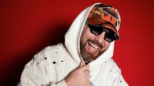Musiker Murda lacht breit mit Kapuzenpulli, Kappi und Sonnenbrille