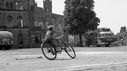 Ein Junge fährt Fahrrad ca. 1945-1955, im Hintergrund zerstörte Wohnhäuser.