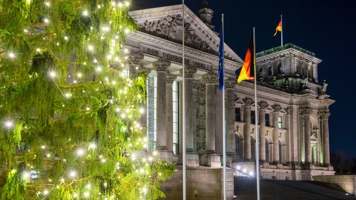 Front des Reichstagsgebäudes in der Dunkelheit mit Deutschland- und Europaflaggen und mit beleuchtetem Weihnachtsbaum im linken Vordergrund