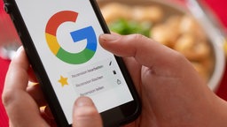 Eine Frau hält vor einem gedeckten Esstisch ein Smartphone mit dem Google-Logo und dem Menüpunkt eine Rezension zu bearbeiten, zu löschen oder zu teilen (Archivfoto vom 17.07.2026)
