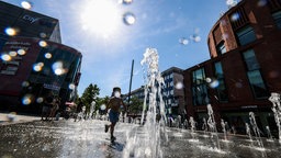 Ein Kind spielt bei am 26.06.2026 bei hochsommerlichen Temperaturen und strahlendem Sonnenschein in den Wasserstrahlen eines begehbaren Brunnens in der Duisburger Innenstadt. 