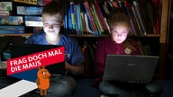 Zwei Kinder sitzen bei Dunkelheit vor einem Regal mit Spielen und Büchern und spielen auf zwei Laptops Computerspiele