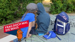 Ein Junge und ein Mädchen sitzen mit einem Ranzen auf einer Tischtennisplatte in einer Grünanlage und schauen gemeinsam auf ein Smartphone