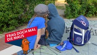Ein Junge und ein Mädchen sitzen mit einem Ranzen auf einer Tischtennisplatte in einer Grünanlage und schauen gemeinsam auf ein Smartphone