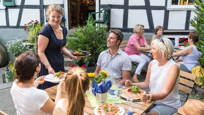Im Außenbereich eines Cafés in einem Fachwerkhaus bringt eine Kellnerin Gästen Essen an den Tisch.