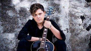 Der Sänger Michael Patrick Kelly schaut in die Kamera und hält eine Gitarre