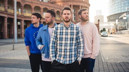 Die 4 Jungs von Revolverheld stehen auf einem Platz und bis auf den Sänger guckt niemand zur Kamera. im Hintergrund sieht man die Sonne über einem Gebäude.
