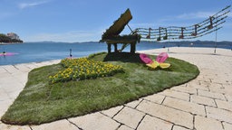 Blumenverzierte Installation eines Flügels auf einer begrünten Insel an einer Uferpromenade. Gelbe Blüten bilden eine Klaviatur, darüber schwebt ein aus Pflanzen gestaltetes Notensystem vor blauem Meer und Himmel.