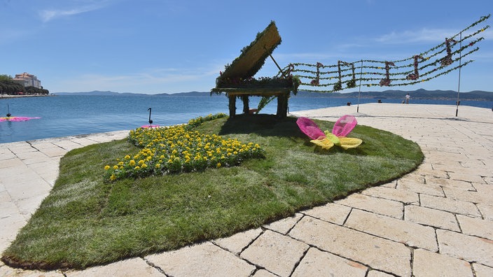 Blumenverzierte Installation eines Flügels auf einer begrünten Insel an einer Uferpromenade. Gelbe Blüten bilden eine Klaviatur, darüber schwebt ein aus Pflanzen gestaltetes Notensystem vor blauem Meer und Himmel.
