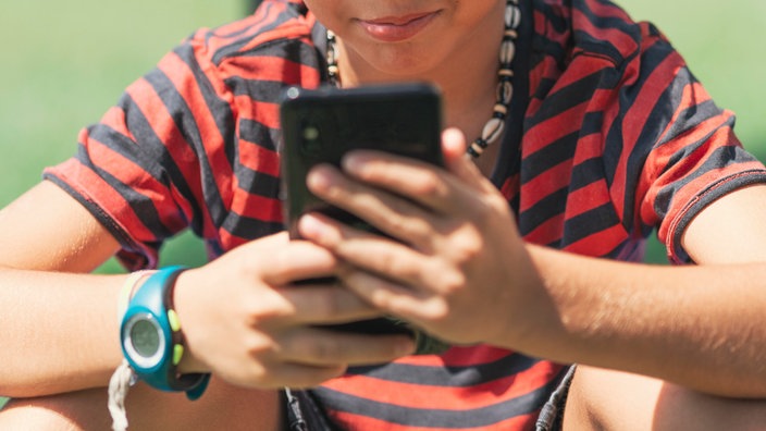 Ein Junge sitzt 2020 in einem Park auf der Wiese und schaut auf ein Handy.