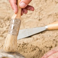 Eine Person hält einen Pinsel und legt einen Gegenstand im Sand frei (Symbolbild).