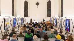 Menschen sitzen umgeben von Bildern bei der Ausstellungseröffnung "Kirchen als Vierte Orte".