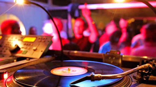 Der Plattenspieler (Turntables) einer Musikanlage, im Hintergrund tanzende Menschen.