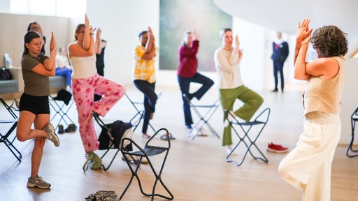 27.07.2026, Nordrhein-Westfalen, Düsseldorf: Unter der Anleitung von Yogalehrerin Simone Hoffmann (r) praktizieren Teilnehmer einer Führung im Museum Kunstpalast an verschiedenen Stationen Yogaübungen. 