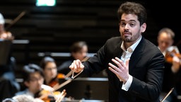 Lahav Shani dirigiert Musiker und Musikerinnen eines Orchesters.