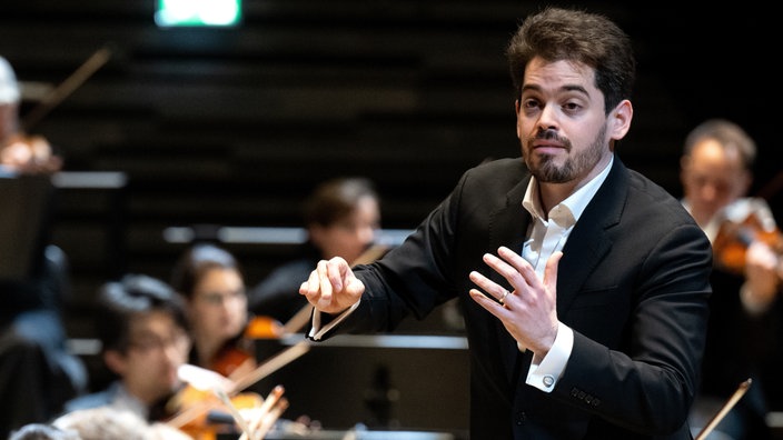 Lahav Shani dirigiert Musiker und Musikerinnen eines Orchesters.