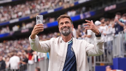 Jürgen Klopp gestikuliert lachend mit beiden Händen und einen Smartphone zum Publikum.