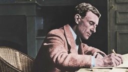 Maurice Ravel in Korbstuhl an Schreibtisch sitzend