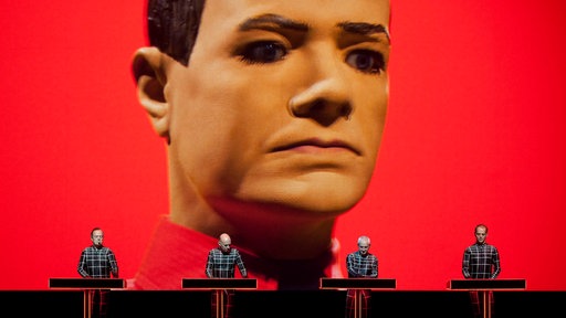Mitglieder von Kraftwerk an elektronischen Instrumenten, im Hintergrund die Projektion eines computer-modellierten Kopfes auf rotem Hintergrund.
