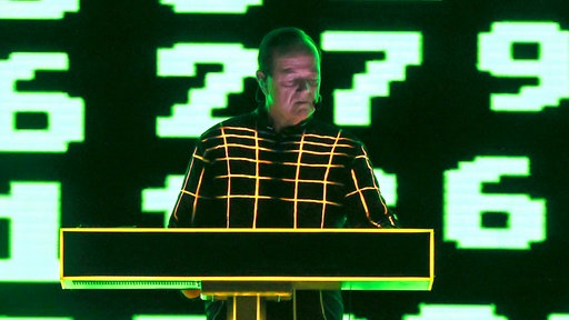 Ralf Hütter bei einem Konzert von Kraftwerk 2025 in einem iluminierten Anzug hinter einem Keyboard.