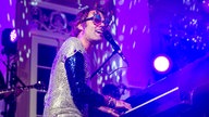 Bastian Korn sitzt mit Glitzeroutfit und einer Spaßbrille mit zwei Gitarren am Klavier