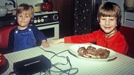 80er-Jahre: Zwei Kinder sitzen an einem Küchentisch und haben Kopfhörer auf, die mit einem Kassettenrekorder verbunden sind.