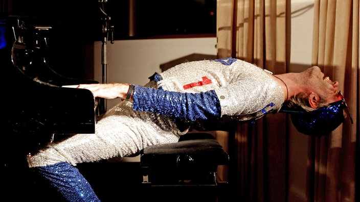 Musiker mit Glitzeroutfit am Flügel akrobatisch nach hinten gebeugt und dabei klavierspielend