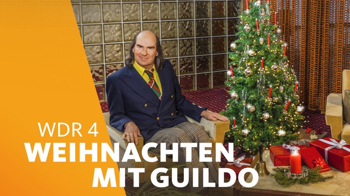 Guildo sitzt auf einem Sessel, neben ihm ein festlich geschmückter Weihnachtsbaum, vor ihm ein Tisch mit Kerze und Geschenken