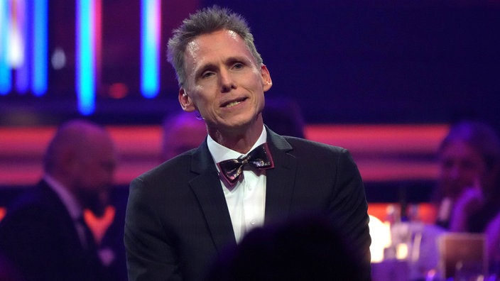 Frank Busemann bei der Sportler des Jahres Gala 2025: Er trägt einen Anzug mit Fliege, der Hintergrund ist blau und rot beleuchtet.
