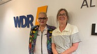 Hella von Sinnen mit Ute Schneider bei WDR 4. Hella von Sinnen trägt eine bunt gemusterte Jacke, Ute Schneider ein weißes, gestreiftes Oberteil. Sie stehen vor einer weißen Wand mit großem WDR 4 Logo.