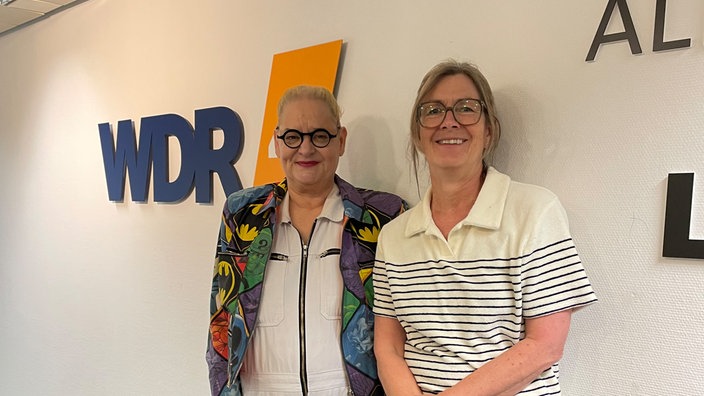 Hella von Sinnen mit Ute Schneider bei WDR 4. Hella von Sinnen trägt eine bunt gemusterte Jacke, Ute Schneider ein weißes, gestreiftes Oberteil. Sie stehen vor einer weißen Wand mit großem WDR 4 Logo.