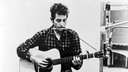Bob Dylan mit einer Mundharmonika um den Hals und einer Gitarre in den Armen (1941).