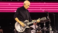 Der Musiker Midge Ure steht mit einer Gitarre auf der Bühne