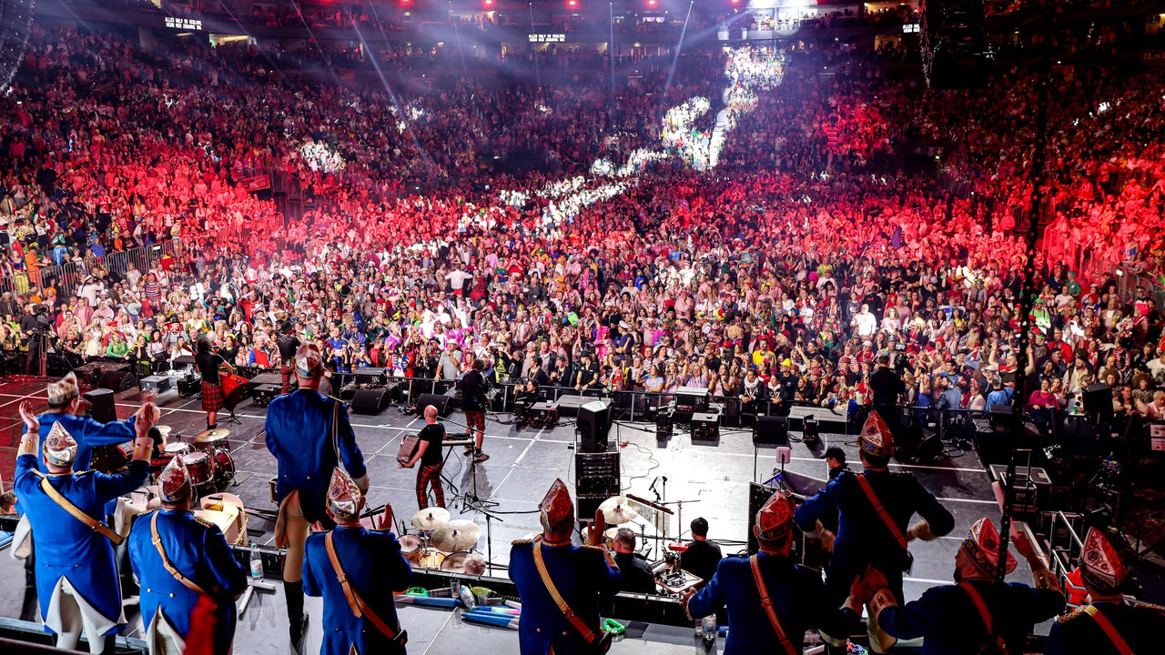Lachende Köln Arena 2026: Ein Blick vom Oberrang hinter der Bühne. Der Elferrat steht auf Stühlen und tanzt hinter der Band mit
