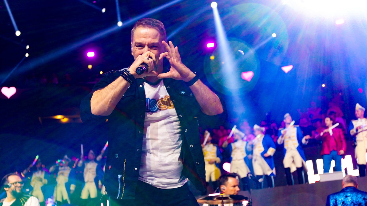 Lachende Köln Arena 2026: Die Band Höhner steht auf der Bühne. Der Sänger schaut in die Kamera und formt mit den Händen ein Herz