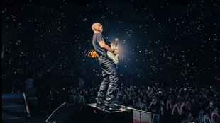 Eros Ramazzotti steht mit einer Gitarre auf der Bühne. Im Hintergrund sieht man im Dunklen das Publikum. 