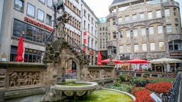 Oben auf dem Brunnen steht Schneidersfraufigur mit einer Laterne in der Hand, etwas weiter unten rechts und links von ihr befinden sich Heinzelmännchenfiguren. Aus der Brunnenschale in der Mitte fließt Wasser in das Brunnenbecken unten.