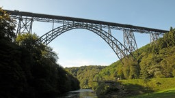 Die Müngstener Brücke zwischen Remscheid und Solingen: Eine stählerne Bogenbrücke vor blauem Himmel. Darunter fließt die Wupper, rechts und links sieht man Bäume und Grünflächen.