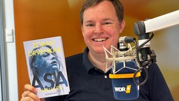 Mike Altwicker sitzt im WDR 4 Studio mit dem Buch "Asa" von Zoran Drvenkar in der einen Hand und einem Mikrofon vor sich