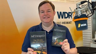 Das Buchcover "Die Straße der Knochen" von Val McDermid wird von Mike Altwicker im WDR 4-Studio in die Kamera gehalten