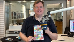 Mike Altwicker steht im WDR 4 Studio am Mikrofon und hält das Buch "Guten Morgen, schönes Wetter heute" von Tanja Kokoska hoch. 