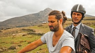 Filmszene aus "Highway to Hellas" (2015): Jörg Geissner (Christoph Maria Herbst) fährt mit Aktentasche unterm Arm bei Panos (Adam Bousdoukos) auf dem Motorrad mit