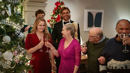 Eine Szene des Films "A Merry Little Ex-Mas": Die Familie steht festlich gekleidet im weihnachtlichen geschmückten Wohnzimmer und stößt mit Sekt an. 