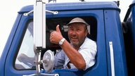 Manfred Krug in der Rolle des Truckers Franz Meersdonk während der Dreharbeiten zu "Auf Achse" in einem blauen Lastwagen. Er sitzt auf dem Fahrersitzt und lehnt sich aus dem Fenster, lächelt in die Kamera und streckt einen Daumen nach oben. 