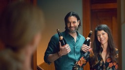 Eine Szene des Films "Die Nachbarn von oben": Die Schauspieler Maximilian Simonischek und Sarah Spale stehen als die neuen Nachbarn mit zwei Sektflaschen vor der Wohnungstür und lächeln breit