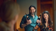 Eine Szene des Films "Die Nachbarn von oben": Die Schauspieler Maximilian Simonischek und Sarah Spale stehen als die neuen Nachbarn mit zwei Sektflaschen vor der Wohnungstür und lächeln breit