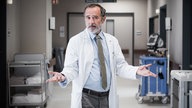 Eine Szene aus der Serie "Doktor Ballouz": Dr. Amin Ballouz (Merab Ninidze) im Arztkittel mit ausgestreckten Armen im Gang eines Krankenhauses