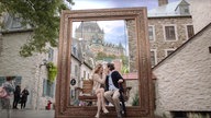 Szene aus dem Film "French Girl": Sophie, gespielt von Evelyne Brochu, und Gordon, gespielt von Zach Braff, sitzen auf einer Bank, die als Fotospot vor einem übergroßen Bilderrahmen aufgestellt ist. Sie nähern sich zum Kuss, ihre Hand ist an seiner Wange.