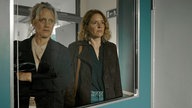 Szene aus der Thriller-Serie "Hundertdreizehn": Riccarda (gespielt von Anna Schudt, links) und Caro (gespielt Patricia Aulitzky) müssen ihren toten Partner identifizieren. Sie schauen durch ein Fenster in einer türkisen Tür.